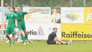 Ein Fußballspieler in einem grünen Trikot feiert ein Tor, während ein anderer Spieler in Schwarz auf dem Boden sitzt, beide vor einem Netz mit Werbung.