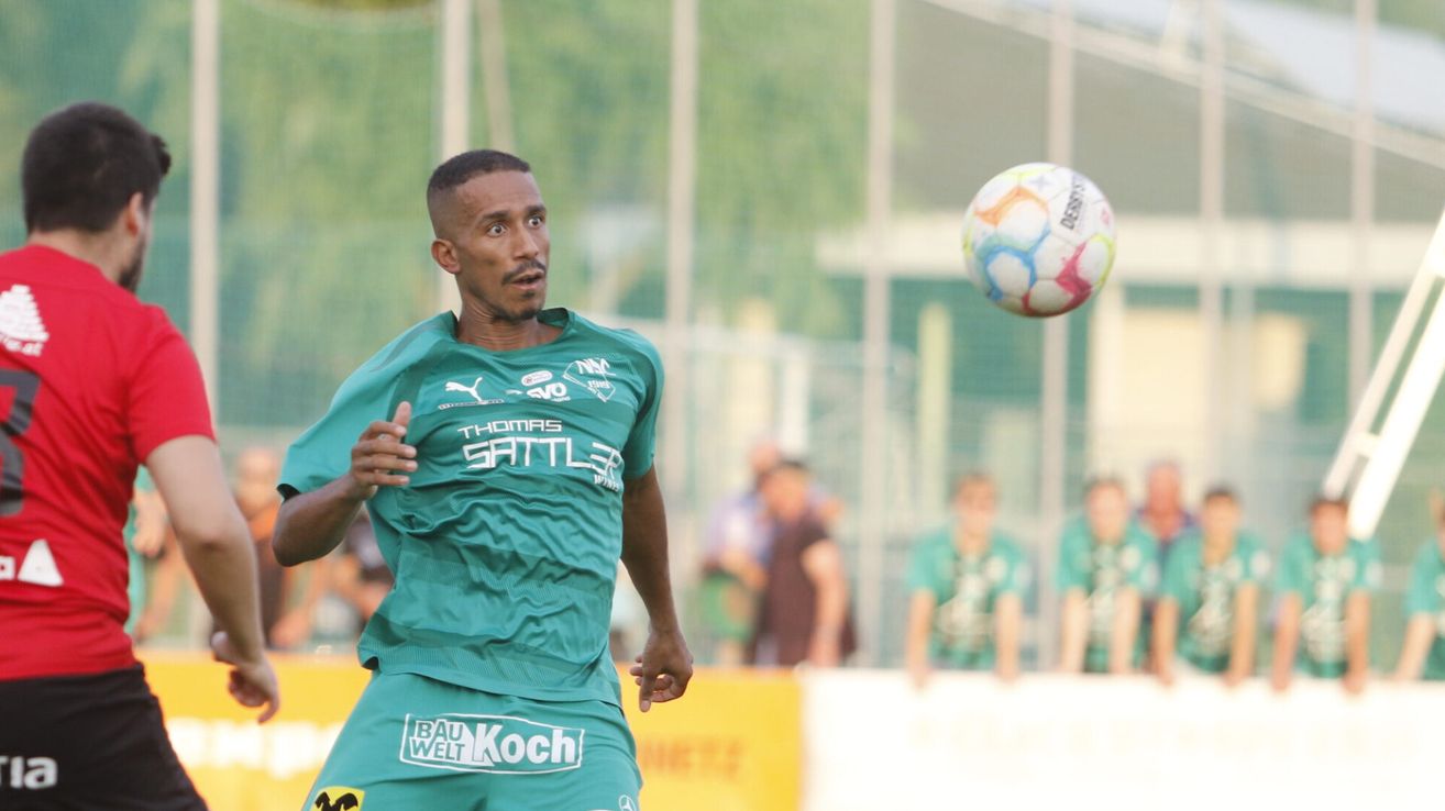 Ein Fußballspieler in einem grünen Trikot mit dem Wort Thomas Satteler darauf läuft auf einen bunten Fußball in der Luft zu.