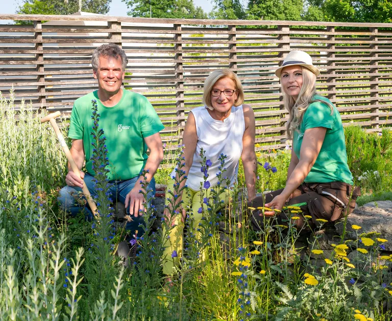 Drei Menschen lächeln und posieren für ein Foto in einem Garten mit verschiedenen Pflanzen und Blumen. Der Mann hält eine Schaufel, und die Frau trägt einen Hut.