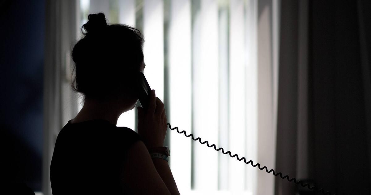 Eine Silhouette einer Frau, die ein Telefon an ihr Ohr hält, möglicherweise in einem Anruf. Sie steht vor einem Fenster mit Jalousien.