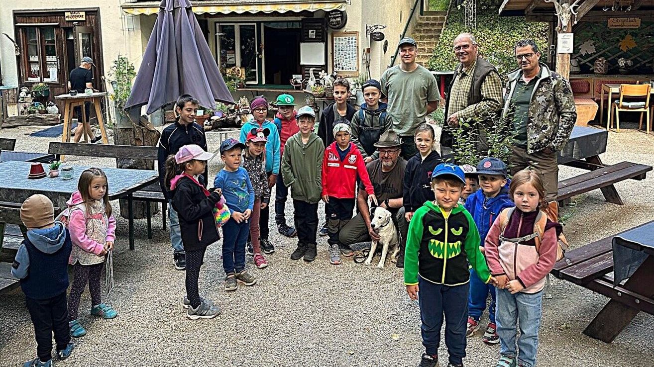 A group of children and adults stand outside a building with a garden, some wearing hats and sneakers, and a man kneeling with a dog.