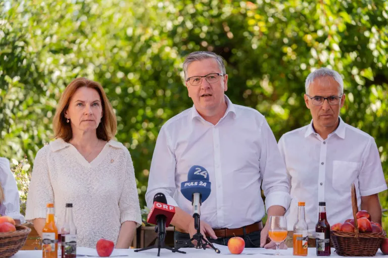 Three people are seated at a table with microphones and bottles. A man in the middle is speaking into a microphone. Behind them, there are trees and a blurred background.