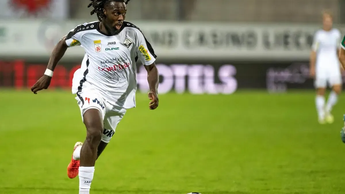 Ein Fußballspieler in einem weißen Trikot mit roten Akzenten läuft auf dem Feld und konzentriert sich auf den Ball vor ihm. Der Hintergrund ist unscharf mit Werbung für Hotels und Casinos.