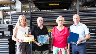 Vier Personen stehen vor einem Lager mit Metallregalen und Rohren, lächeln und halten Gegenstände wie einen Kuchen und eine blaue Mappe.