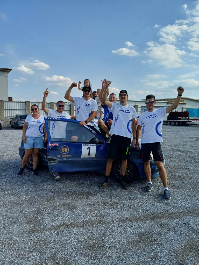 Eine Gruppe von Menschen in weißen Shirts und Shorts posiert für ein Foto vor einem blauen Auto mit der Nummer 1. Sie alle lächeln und heben ihre Hände. Hinter ihnen sind einige Autos geparkt und ein Gebäude mit einem Zaun.