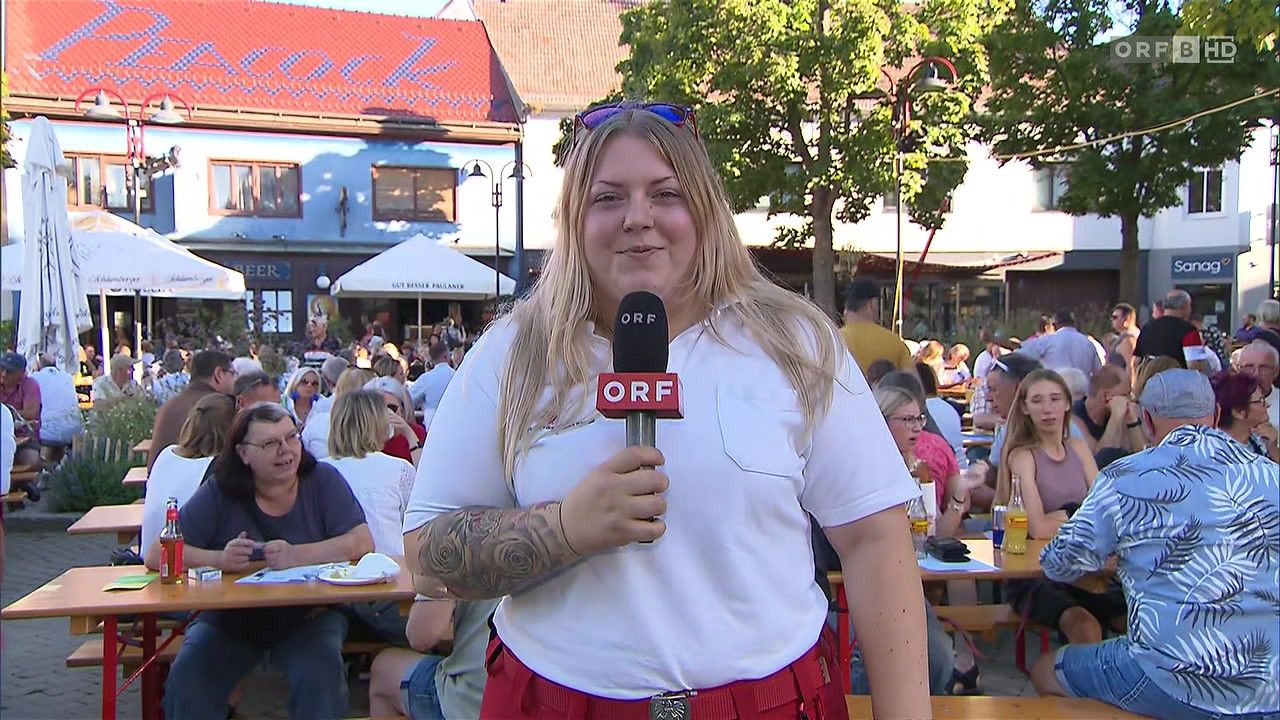 A woman with tattoos on her arm holds a microphone with ORF logo, standing in a crowded outdoor area with people sitting at tables. Behind her, a building with a red roof and a white canopy is visible.