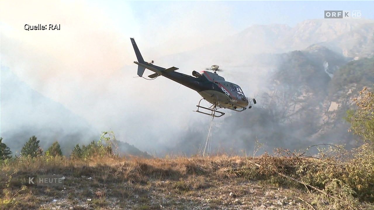 Ein Hubschrauber mit dem Wort Rock an der Seite fliegt über einem Grasfeld, möglicherweise im Einsatz bei Löscharbeiten.