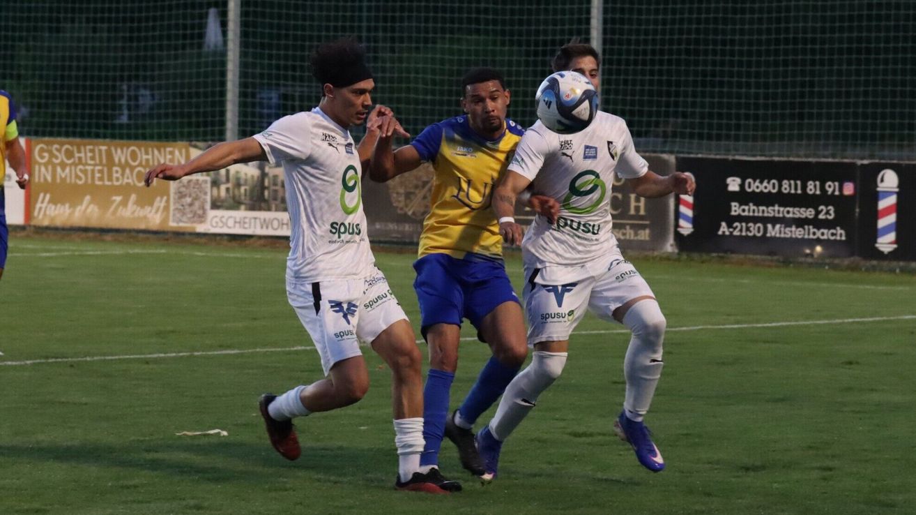 Three soccer players are in action on a field. One player is attempting to head the ball while two others try to block him.