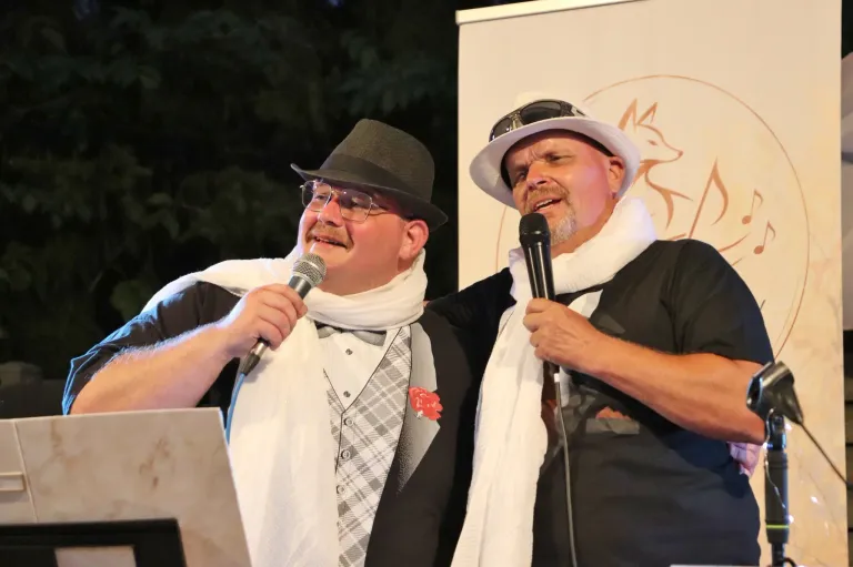 Two men dressed in formal attire, one with a bow tie and a red flower, and the other with a white scarf, are standing and holding microphones. They are smiling and seem to be performing on stage.