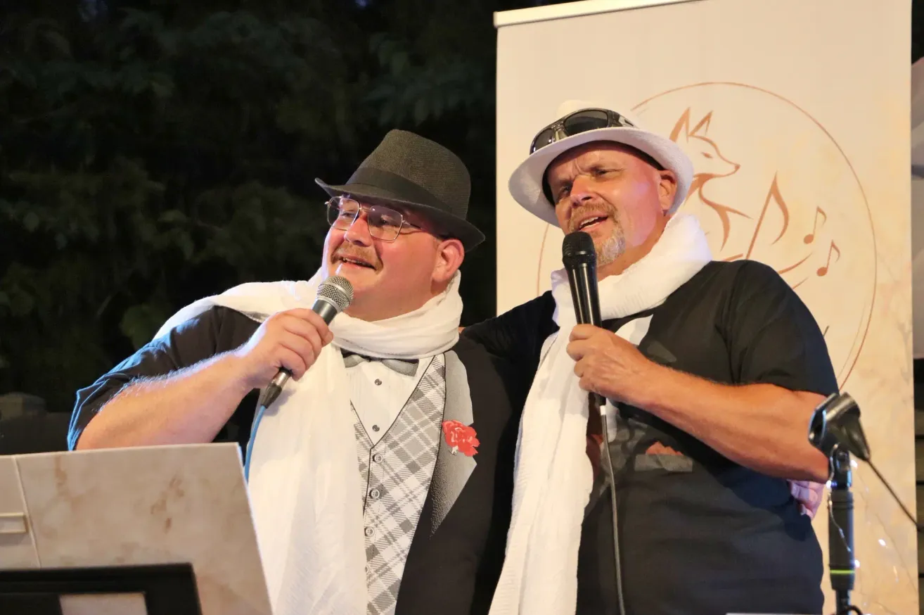 Two men dressed in formal attire, one with a bow tie and a red flower, and the other with a white scarf, are standing and holding microphones. They are smiling and seem to be performing on stage.