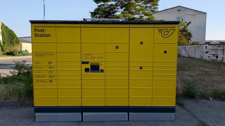 A yellow locker system with various size compartments labeled M, L, S, and XL. The lockers are arranged in a row with a payment machine in the middle.