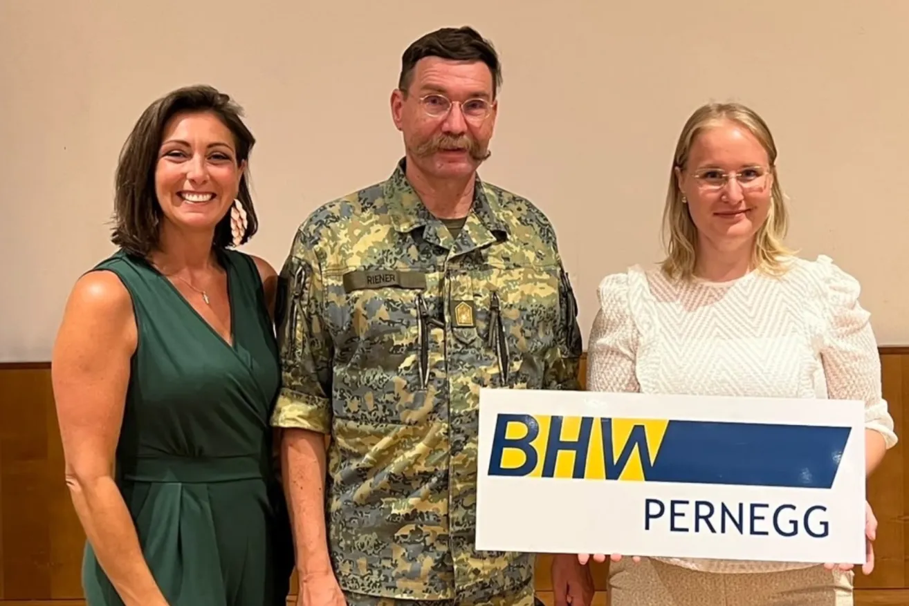 A man in a military uniform stands between two women, holding a sign that reads 'BHW Perne'. The man has a mustache and glasses. The woman on the left wears a green dress, and the woman on the right wears a white blouse. They all smile for the camera.