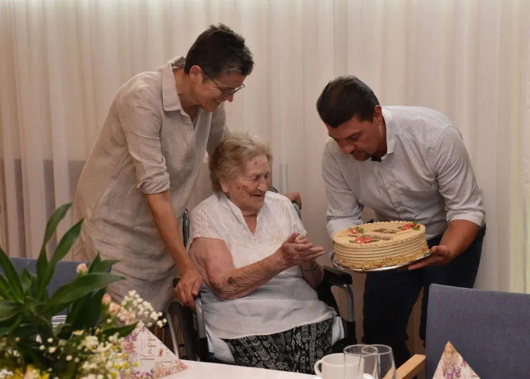 An elderly woman in a wheelchair is celebrating her birthday with two people, one holding a cake and the other smiling.