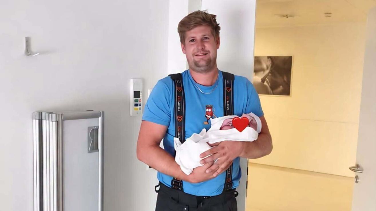 A man in a blue shirt and suspenders is holding a newborn baby in his arms. The baby has a red heart on its blanket.
