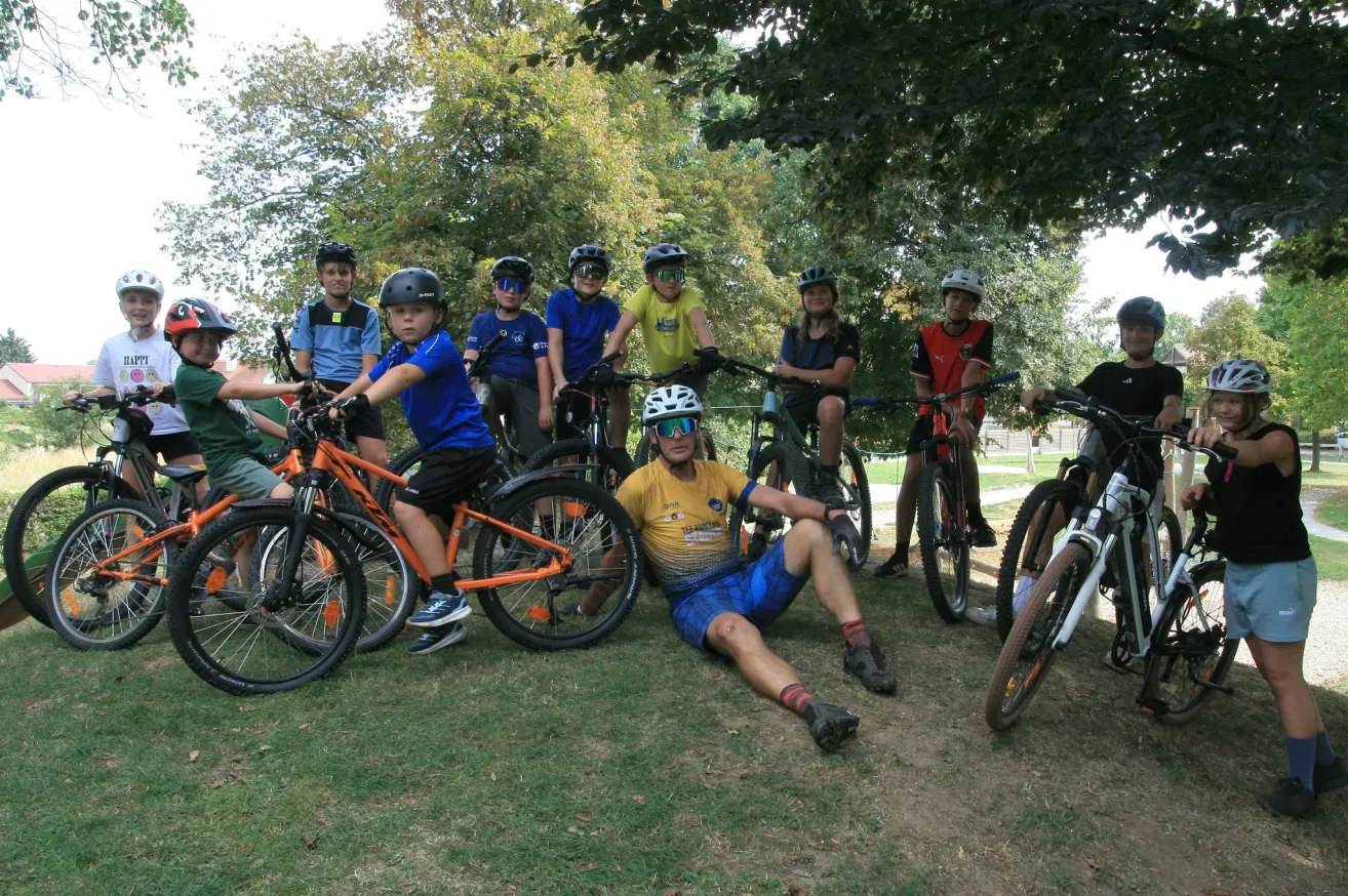 A group of children and a man are posing for a photo with their bikes in a park. The children are wearing helmets and cycling gear.