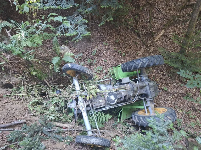 An overturned tractor is stuck in a forest with its front wheels in the air and the back wheels buried in leaves.