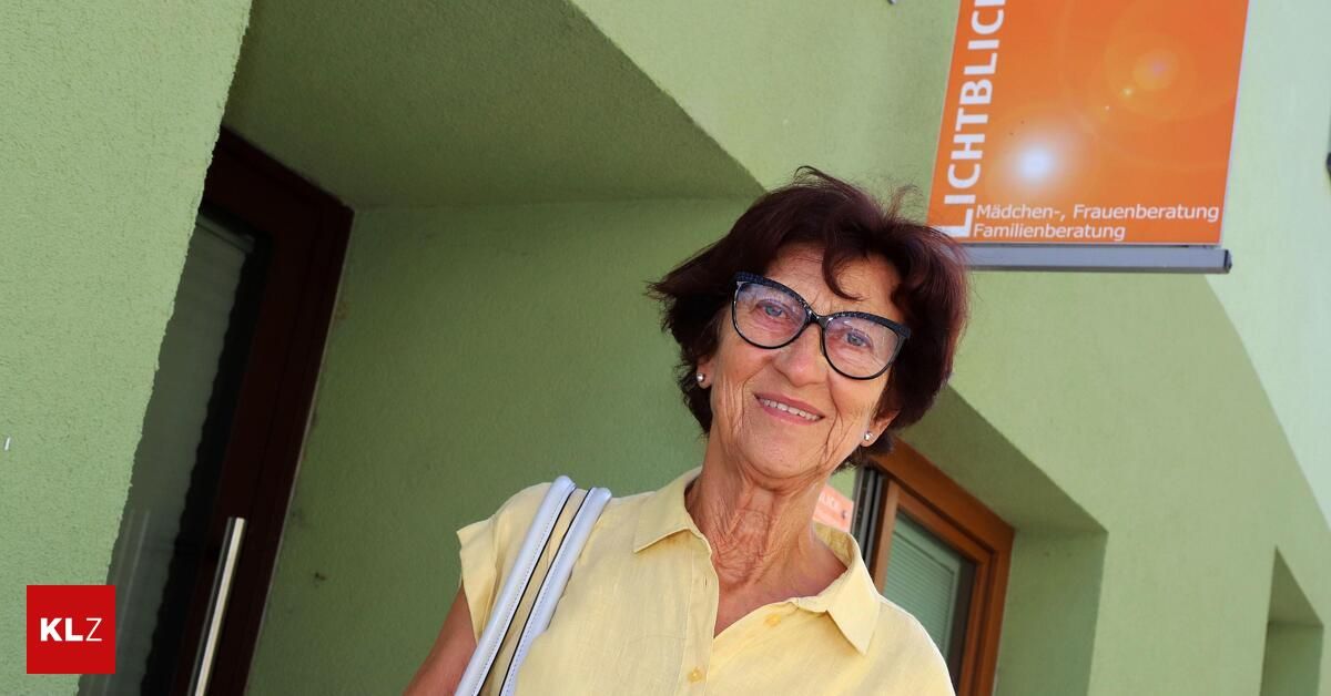 An elderly woman with short hair and glasses is smiling in front of a green wall with a sign that reads 'Lichtblick'.