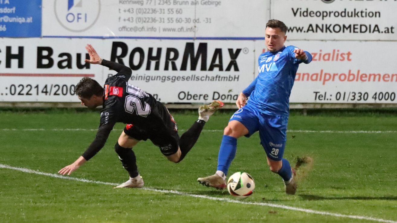 Zwei Fußballspieler in Aktion auf einem Feld. Ein Spieler in Schwarz mit der Nummer 62 auf dem Rücken fällt, während der andere Spieler in Blau mit der Nummer 20 auf den Ball zuläuft.