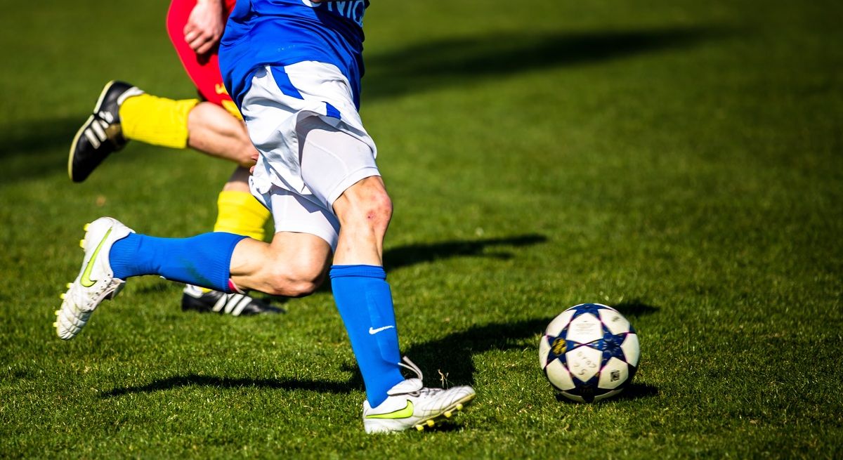 A soccer player in a blue jersey with white shorts is kicking a soccer ball on a green field.