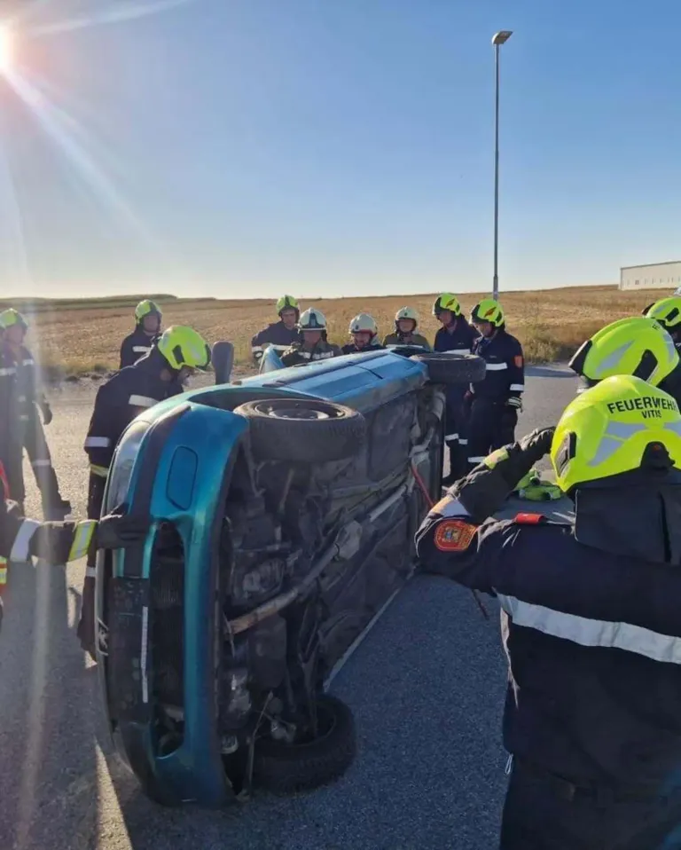 Emergency responders in yellow helmets work on a flipped blue car on the side of a road.