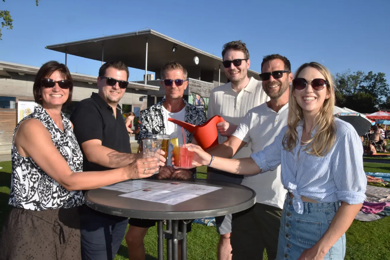 A group of people, all wearing sunglasses, are gathered around a table with drinks. They are smiling and posing for a photo. Behind them, there is a building with a clock on the wall.