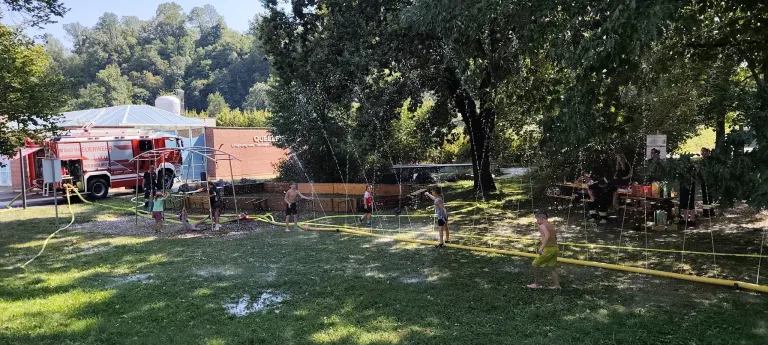 Several children play with water sprinklers in a park, with a building labeled 'QUELL' in the background.