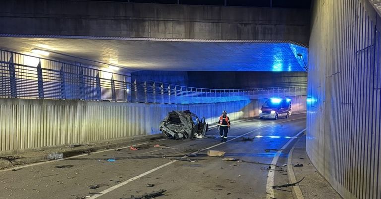Ein Rettungsfahrzeug ist am Straßenrand geparkt, ein Beamter steht neben einem demoliert aussehenden Auto. Die Szene ist von blauen Lichtern beleuchtet, und die Straße ist von einer Betonbarriere umgeben.