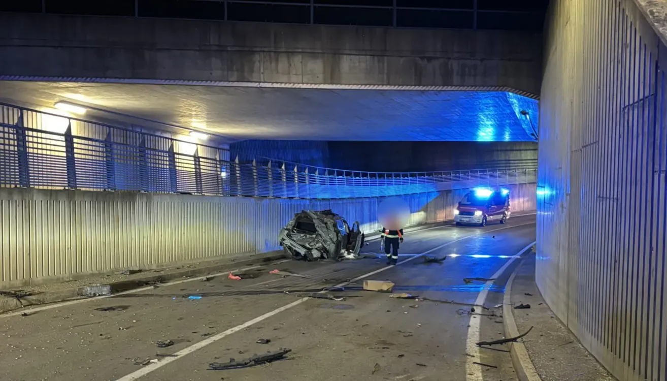 Ein Rettungsfahrzeug mit eingeschalteten Lichtern steht am Straßenrand, in der Nähe eines demolierteren Autos. Eine Person in einer reflektierenden Weste geht auf das demolierte Auto zu.