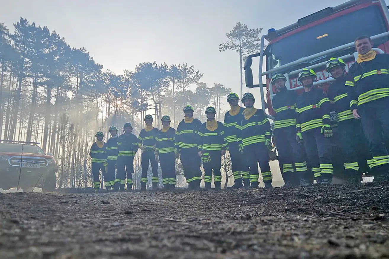 A group of firefighters in uniform stand in a row on a dirt road, with a fire truck behind them. They are wearing helmets and gloves.