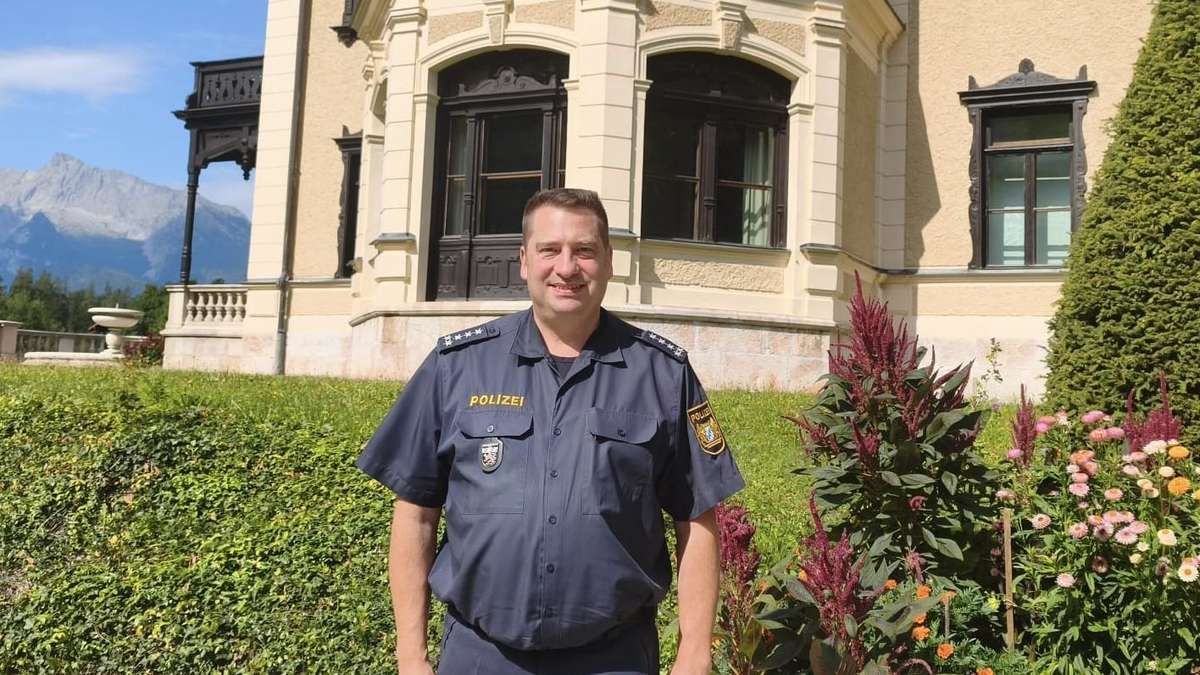A man in a police uniform stands in front of a building with a large window and a mountain in the background.