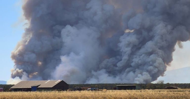 A large plume of smoke rises from a fire in a rural area, with a building visible in the foreground.