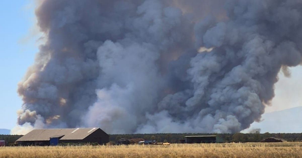 A large plume of smoke rises from a fire in a rural area, with a building visible in the foreground.