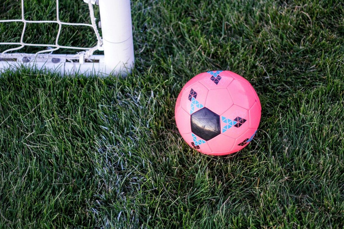 A pink soccer ball with blue diamond patterns lies on a well-maintained grassy field near a white goalpost.
