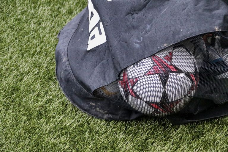 Ein Fußball mit schwarz-weißem Design ist teilweise von einer schwarzen Tasche mit einem weißen Logo auf der Seite bedeckt und liegt auf einem grünen Feld.