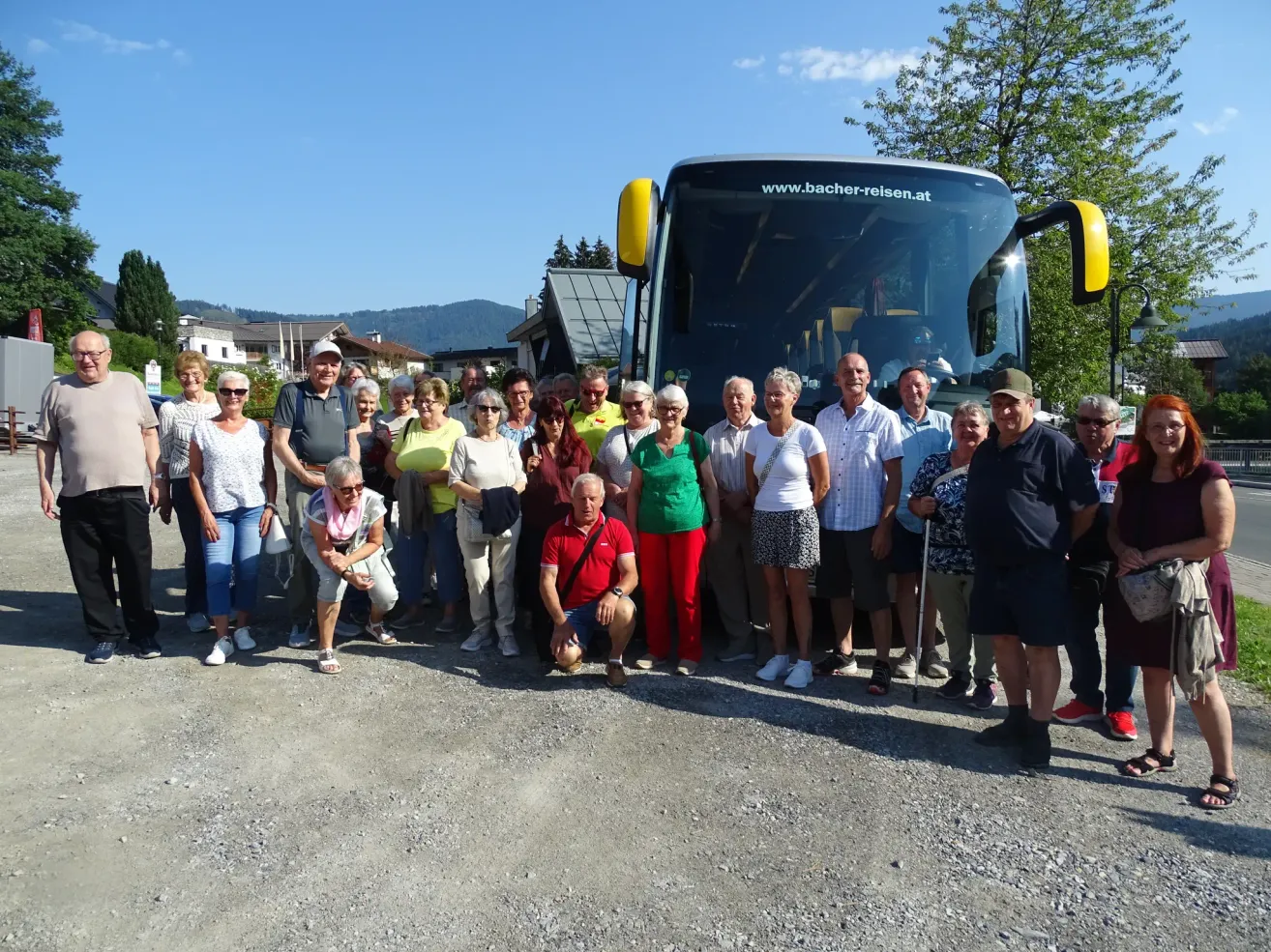 A group of people, possibly tourists, are posing for a photo in front of a bus with the website www.bacher-reisen.at on the front.