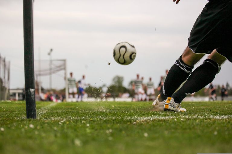 A soccer player kicks a ball in mid-air, with a blurred background of players and spectators on a grassy field.