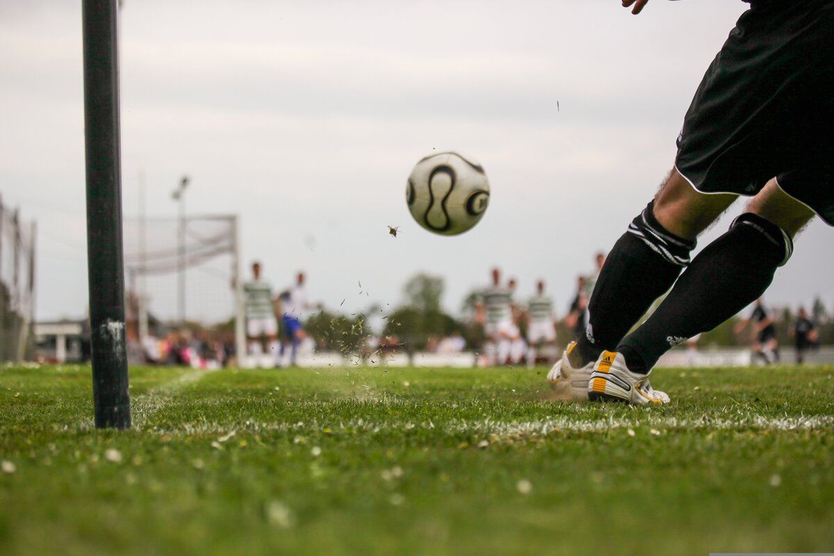 A soccer player kicks a ball in mid-air, with a blurred background of players and spectators on a grassy field.