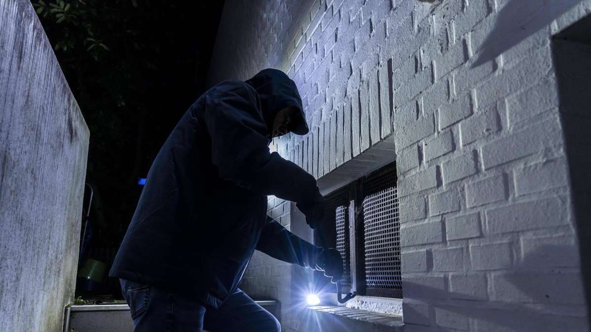 A man in a dark jacket and gloves is using a tool to pry open a window at night.