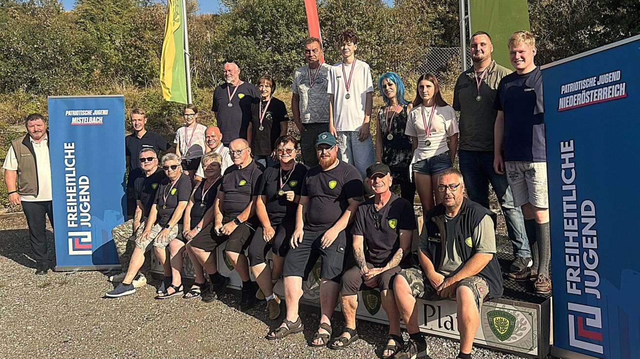 A group of people wearing medals and black t-shirts are posing for a photo on a gravel surface. They are sitting on a white bench with a green banner in the background.