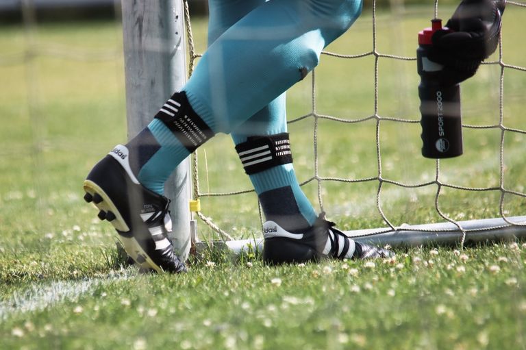 A close-up view of a soccer player's legs in blue and black Adidas gear, standing near a soccer goal.
