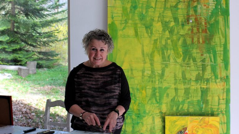 An older woman with gray hair smiles and poses for a photo in front of a vibrant green and yellow abstract painting.