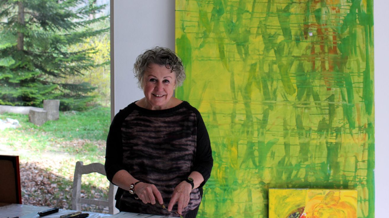 An older woman with gray hair smiles and poses for a photo in front of a vibrant green and yellow abstract painting.
