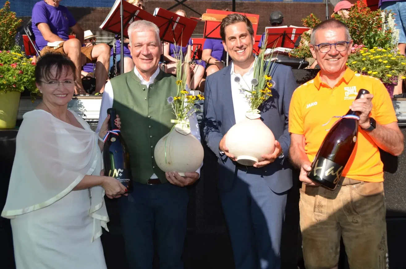 Four people stand together holding large vases with flowers, two of them holding bottles of wine. Behind them, a band is playing with a crowd watching.