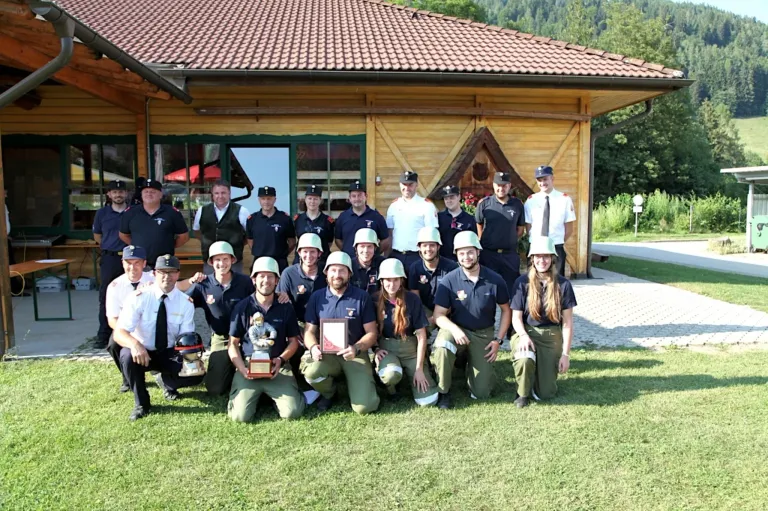 A group of firefighters in uniform pose for a photo in front of a wooden building with a red roof. Some are kneeling, others standing, and they all wear helmets. A man in the middle holds a trophy.