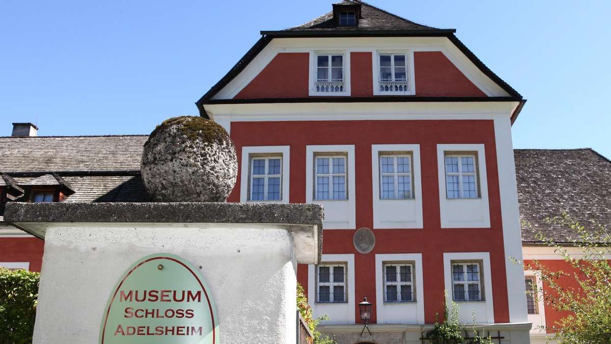 A red and white building with a large stone sphere in front, labeled 'Museum Schloss Adelsheim'. The building has multiple windows and a lamp on the side.