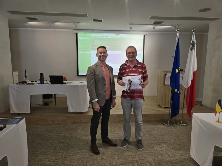 Two men stand in a conference room, one holding a certificate. Behind them, a projector screen displays a presentation. A podium with flags is on the right, and a table with a laptop is on the left.
