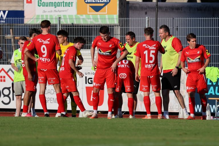 A group of soccer players in red uniforms are standing on the field, possibly discussing the game. The player in the middle is looking down, while the others are looking at him. Behind them is a fence with a banner.
