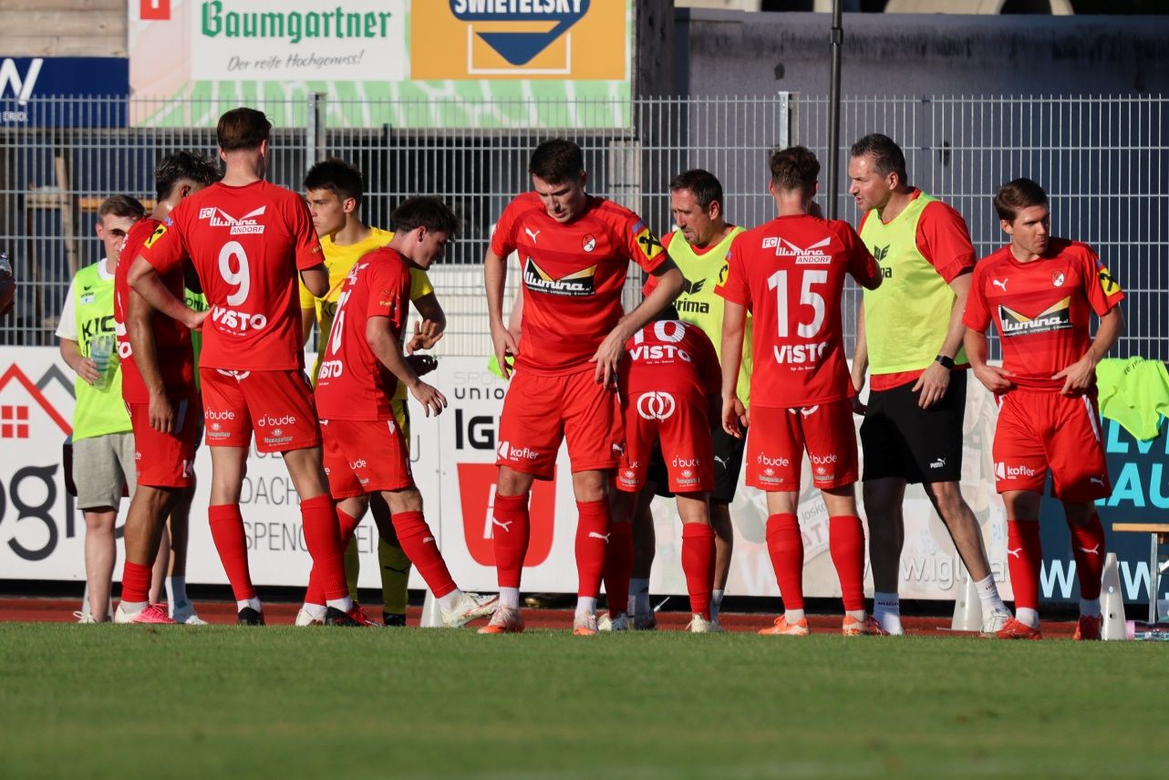 A group of soccer players in red uniforms are standing on the field, possibly discussing the game. The player in the middle is looking down, while the others are looking at him. Behind them is a fence with a banner.