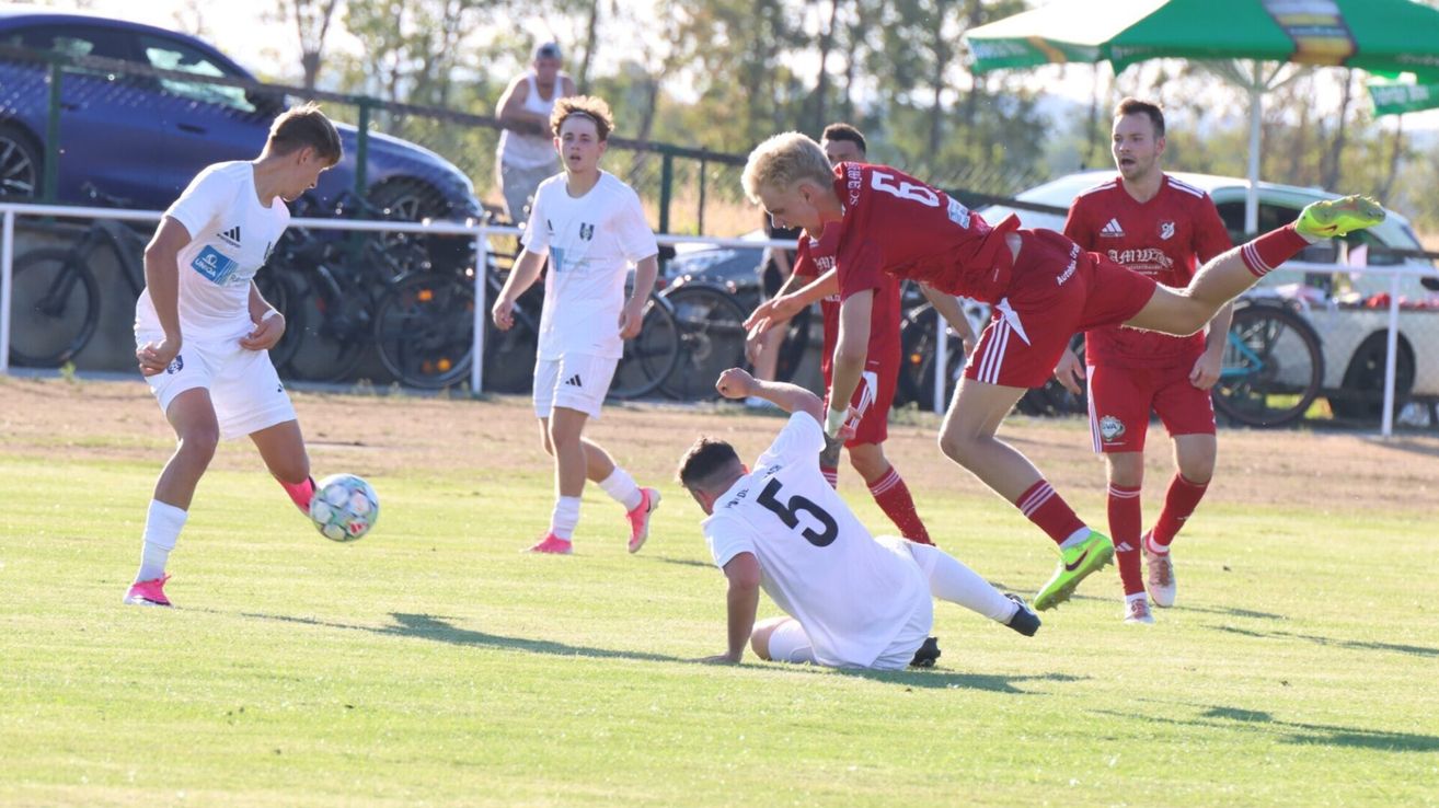 A soccer match with players in red and white uniforms, one player in red is jumping while another in white is on the ground.