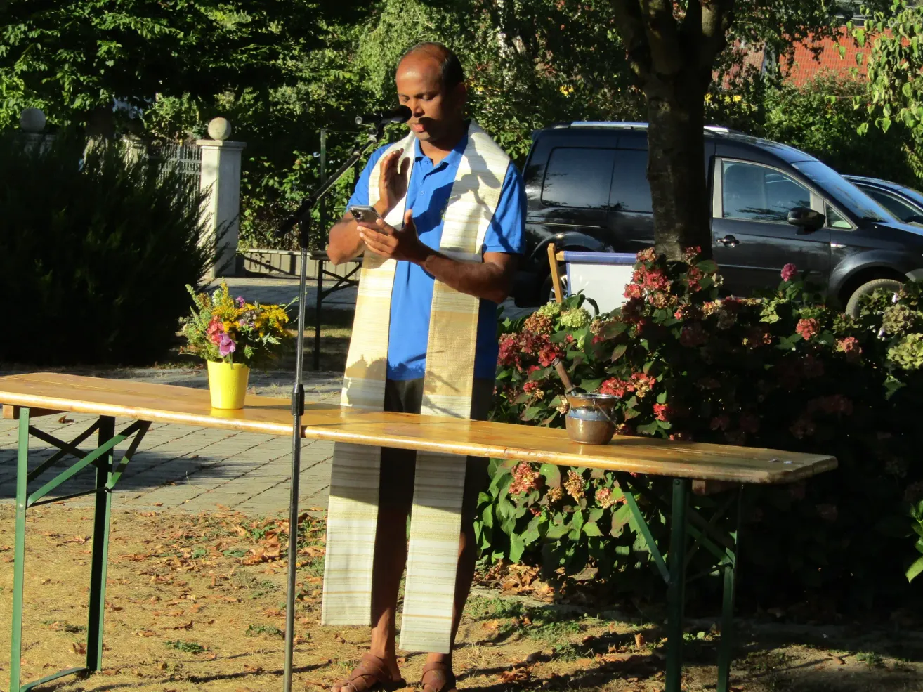 A man wearing a white robe stands behind a table with a microphone, holding a cellphone. Potted flowers and a small pot are on the table. Behind him is a tree, a van, and a fence.